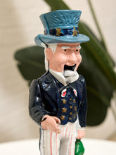 Load image into Gallery viewer, A hand-painted Uncle Sam figurine wearing a blue top hat, dark blue jacket, white pinstriped trousers, and holding a green sack, set against a soft-focus background with a green plant.