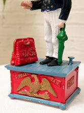 Load image into Gallery viewer, A vintage metal toy bank or collectible box featuring a red body with a gold eagle relief, a blue top, a red suitcase detail, and a green umbrella detail, with a small figurine of a man standing on top.