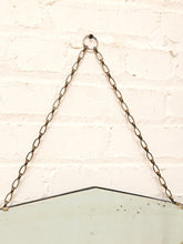 Load image into Gallery viewer, A vintage-style hanging mirror with a triangular frame, suspended by a bronze chain from a ring on a white painted brick wall.