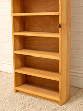 Load image into Gallery viewer, A tall, five-tier wooden bookshelf in a light natural oak finish, standing on a textured beige rug against a white brick wall.