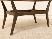 Load image into Gallery viewer, A close-up of a dark brown wooden bench or low table with angled legs and a slatted front, set on a light beige woven rug against a white brick wall.