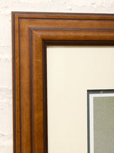 Load image into Gallery viewer, Close-up of a dark brown wooden picture frame with a cream mat board and a portion of a framed print visible against a white brick wall.