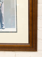 Load image into Gallery viewer, A close-up of a framed art print showing the corner of a dark brown wooden frame with a cream mat, mounted on a white brick wall.
