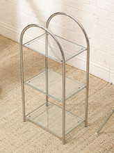 Load image into Gallery viewer, A three-tier glass shelf stand with a polished silver metal frame and arched top handles, set on a beige woven rug against a white brick wall.