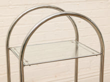 Load image into Gallery viewer, A modern two-tier metal and glass cart with a polished chrome frame and clear glass shelves, set against a light beige brick wall.