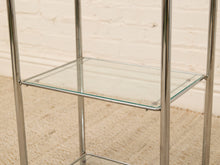Load image into Gallery viewer, A modern two-tier shelving unit with a polished chrome metal frame and clear glass shelves, set against a white brick wall and beige carpet.