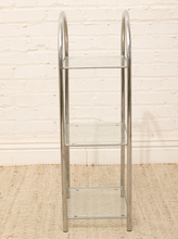 Load image into Gallery viewer, A three-tier shelving unit with a polished chrome metal frame and clear glass shelves, standing on a textured beige rug against a white brick wall.