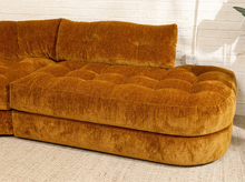 Load image into Gallery viewer, A mid-century modern curved sofa upholstered in a rich mustard-yellow velvet fabric, set against a white brick wall and light-colored carpet.