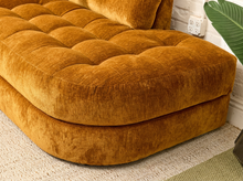 Load image into Gallery viewer, A close-up of a tufted velvet ottoman or sofa cushion in a warm camel-brown color, shown in a styled interior setting with a white brick wall, rug, and green plant leaf.