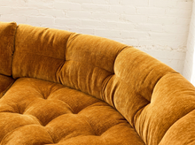Load image into Gallery viewer, Close-up of a tufted velvet sofa in a warm camel or ochre-brown color, set against a white painted brick wall.