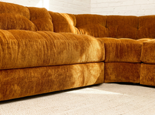 Load image into Gallery viewer, A close-up of a plush tufted sectional sofa upholstered in a warm camel-brown velvet fabric, set against a white brick wall and light beige carpet.