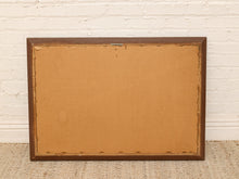 Load image into Gallery viewer, The back of a rectangular wooden picture frame with a brown wood border and tan cardboard backing, leaning against a white brick wall on a woven rug.