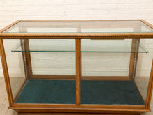 Load image into Gallery viewer, A vintage wooden display case with a glass top and two glass shelves, featuring a dark teal velvet lining, set against a white brick wall background.