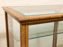 Load image into Gallery viewer, Close-up of a wooden display case with a glass top and clear glass shelves, showing carved wood trim and a warm medium-brown finish against a light brick wall.