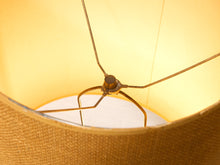 Load image into Gallery viewer, Close-up of the interior of a woven fabric lampshade with a brass metal shade support rod visible against a warm yellow light.
