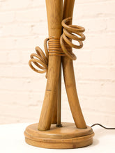Load image into Gallery viewer, A vintage-style table lamp made from natural wood and rattan, featuring a round wooden base and three angled wooden legs wrapped with coiled rattan details, set against a white brick wall background.
