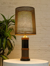 Load image into Gallery viewer, A table lamp with a large cylindrical woven shade, a dark column base, and warm wood-toned bands, shown lit on a white pedestal against a white brick wall with a green plant at the side.