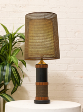 Load image into Gallery viewer, A table lamp with a large woven brown and gold drum shade, a black cylindrical base, and warm wood-toned bands, displayed on a white pedestal against a white brick wall with a green plant on the left.