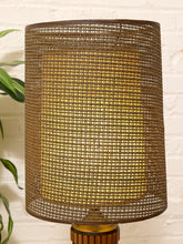 Load image into Gallery viewer, A cylindrical table lamp shade made of a woven natural fiber material, glowing with a warm yellow light from within. The shade is mounted on a wooden base against a white brick wall background with a green plant leaf visible on the left.