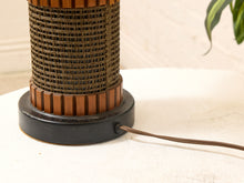 Load image into Gallery viewer, A close-up of a vintage-style table lamp featuring a cylindrical woven brown shade, a black base, and a visible power cord on a light fabric surface.