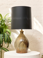 Load image into Gallery viewer, A table lamp with a matte black drum shade and a glazed ceramic base featuring brown and beige marbled patterns, displayed on a white pedestal against a white brick wall with a green plant on the left.