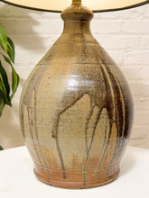 Load image into Gallery viewer, A ceramic table lamp with a rounded bulbous base featuring a brown and beige glaze with dark drips, topped with a cream-colored shade, set against a white brick wall background.