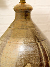 Load image into Gallery viewer, Close-up of a rustic ceramic lamp base with a textured, speckled glaze in earthy brown, beige, and gold tones, featuring a small brass finial at the top against a white brick wall background.
