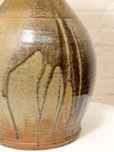 Load image into Gallery viewer, A close-up of a ceramic vase with a rounded body, featuring a glossy brown and beige drip glaze pattern against a light brick wall background.