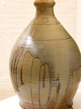 Load image into Gallery viewer, A handmade ceramic vase with a rounded bulbous body, a narrow neck, and a small flared rim. It features a speckled earthy brown and beige glaze with prominent dark brown drip patterns running vertically down the front, set against a light brick wall background.