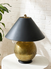 Load image into Gallery viewer, A table lamp with a matte black conical shade and a textured metallic gold spherical base, displayed on a white round table against a white brick wall with a green plant leaf visible on the left.