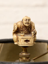 Load image into Gallery viewer, An antique carved ivory or bone figurine of a seated figure with a painted face, mounted on a brass-colored cylindrical base, shown close-up against a softly blurred light background.