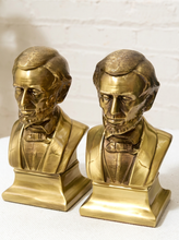 Load image into Gallery viewer, A pair of gold-toned Abraham Lincoln bust bookends on a white textured surface against a white brick wall background.