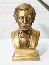 Load image into Gallery viewer, A metallic gold Abraham Lincoln bust sculpture on a matching rectangular base, photographed against a white brick wall background.