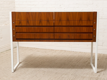 Load image into Gallery viewer, A mid-century modern wooden console table with a warm brown walnut-grain finish and white metal legs, set against a white brick wall and beige carpet.