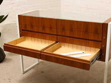 Load image into Gallery viewer, A mid-century modern wooden desk with a glass top, featuring two open drawers with light wood interiors and a white metal frame, set against a white brick wall.