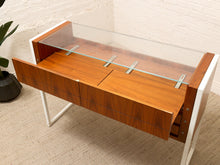 Load image into Gallery viewer, Mid-century modern wooden display cabinet with a warm brown wood grain finish, white side panels and legs, a clear glass top shelf, and a lower drawer pulled open, set against a white brick wall and beige carpet.