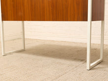 Load image into Gallery viewer, Close-up of a mid-century modern wooden console table with a warm brown wood grain front and white metal legs, set on a beige carpet against a white brick wall.