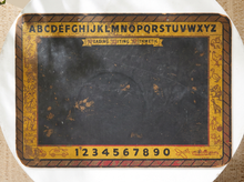 Load image into Gallery viewer, A vintage reading, writing, and arithmetic practice board with a black central writing area and a yellow border printed with alphabet letters, numbers, and small illustrative icons, shown on a white surface.