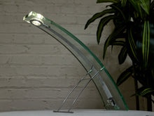 Load image into Gallery viewer, A modern glass table lamp with a curved clear glass shade and a silver metal stand, shown lit against a light brick wall with a green plant in the background.