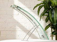 Load image into Gallery viewer, A modern glass table lamp with a curved clear glass shade and slim silver metal stand, set against a white brick wall with a green plant in the background.