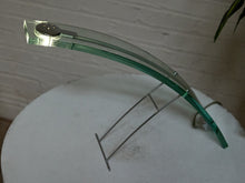 Load image into Gallery viewer, A modern curved glass shelf or console table with a clear glass top, green-tinted glass side panels, and a slim metal support frame, displayed on a round white pedestal against a white brick wall.