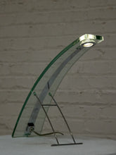 Load image into Gallery viewer, A modern glass table lamp with a curved transparent glass shade and a slim metal stand, shown lit against a white brick wall background.