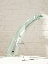 Load image into Gallery viewer, A modern table lamp made of transparent curved glass with a silver metal stand, set against a white brick wall background.