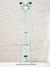 Load image into Gallery viewer, A modernist table lamp with a tall, clear glass column, a polished silver metal base and support structure, and a circular glass shade at the top, set against a white brick wall background.