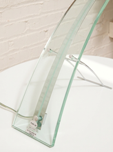 Load image into Gallery viewer, A modern curved glass table lamp with a clear glass shade and a silver metal support stand, resting on a white surface against a white brick wall.
