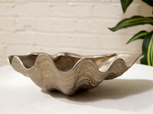 Load image into Gallery viewer, A metallic silver clamshell-shaped bowl with a wavy rim, displayed on a white surface against a white brick wall background with a green plant leaf in the corner.