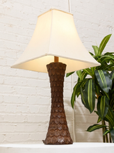 Load image into Gallery viewer, A table lamp with a textured, dark brown carved base and a warm cream-colored fabric shade, set against a white brick wall with a green plant in the background.