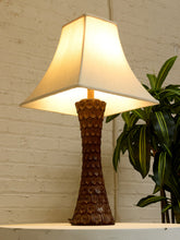 Load image into Gallery viewer, A table lamp with a textured dark brown ceramic base and a warm white fabric shade, shown lit on a white surface against a white brick wall with a green plant in the background.