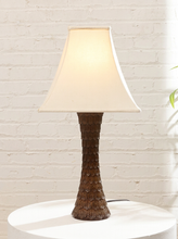 Load image into Gallery viewer, A table lamp with a textured brown base and a warm off-white conical shade, illuminated against a white brick wall background.