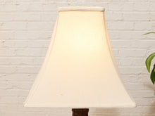 Load image into Gallery viewer, A classic tapered fabric lampshade in a warm cream or off-white shade, glowing with a soft warm light. It is set against a white painted brick wall with a green plant leaf visible on the right side.